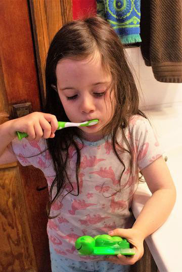 young girl with long brown hair wearing pajamas with pink dinosaurs brushing teeth while looking down at the green plastic two minute turtle timer